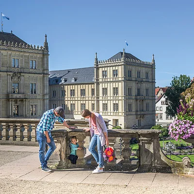 Familienausflug bei Schloss Ehrenburg (Coburg, Coburg.Rennsteig) Eine bunt gekleidete Familie ist vor einem Schloss, welches aus mehreren Bauten besteht, zu sehen. Im Hintergrund ist ein Schlossgarten erkennbar.