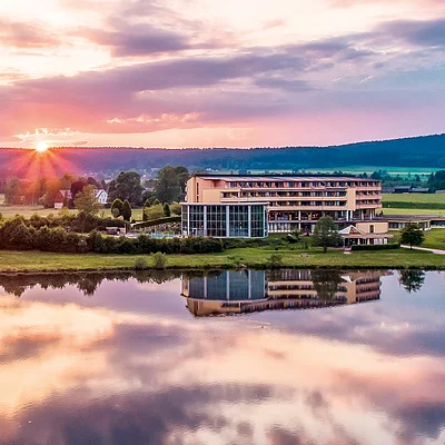 Ein modernes Gebäude am Ufer eines Sees bei Sonnenuntergang, umgeben von grünen Feldern und Bäumen.