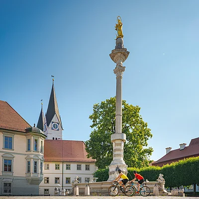 Radler am historischen Residenzplatz (Eichstätt, Naturpark Altmühltal) Zwei Radfahrer fahren an einer Mariensäule vorbei, im Hintergrund eine Kirche und Gebäude.