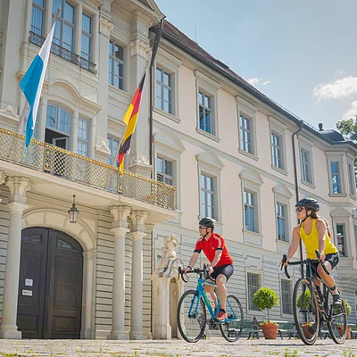 Zwei Personen fahren mit Fahrrädern vor einem historischen Gebäude mit Flaggen.