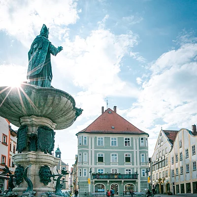 Brunnen mit Statue auf einem Platz, umgeben von historischen Gebäuden und Menschen, die spazieren gehen.