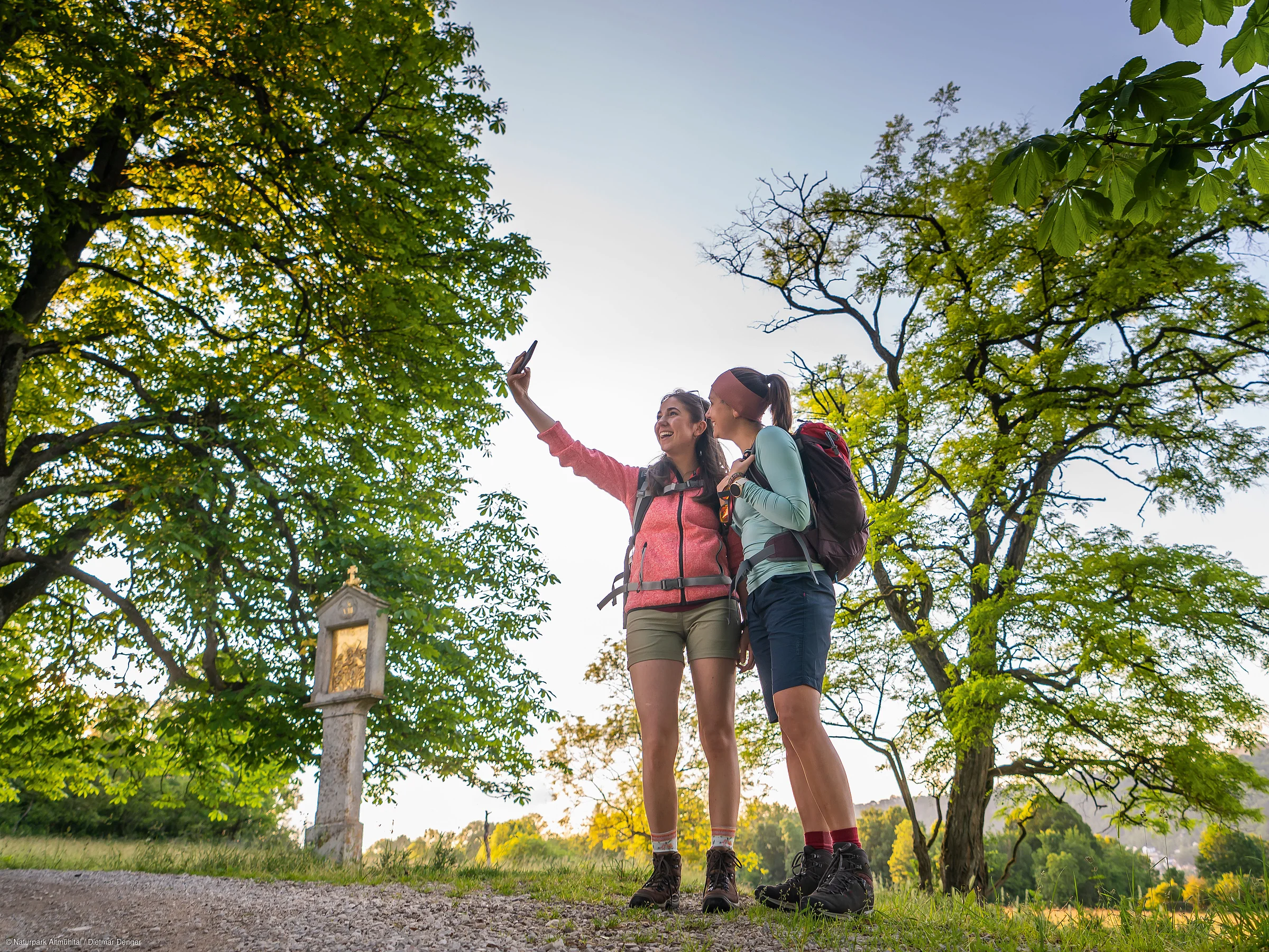 Zwei Personen mit Rucksäcken machen ein Selfie in einer grünen, bewaldeten Landschaft.