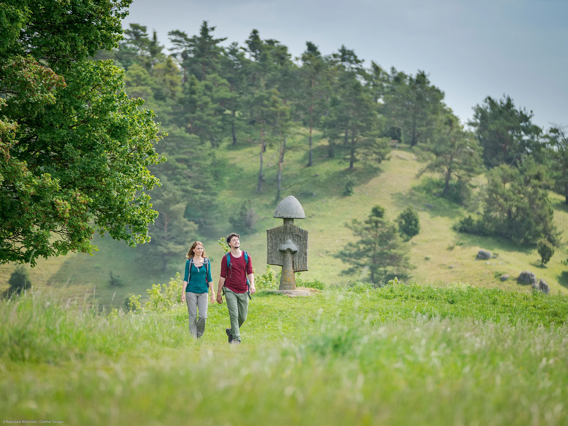 Zwei Personen wandern auf einem grasbewachsenen Weg, im Hintergrund Skulpturen, Bäume und Hügel.