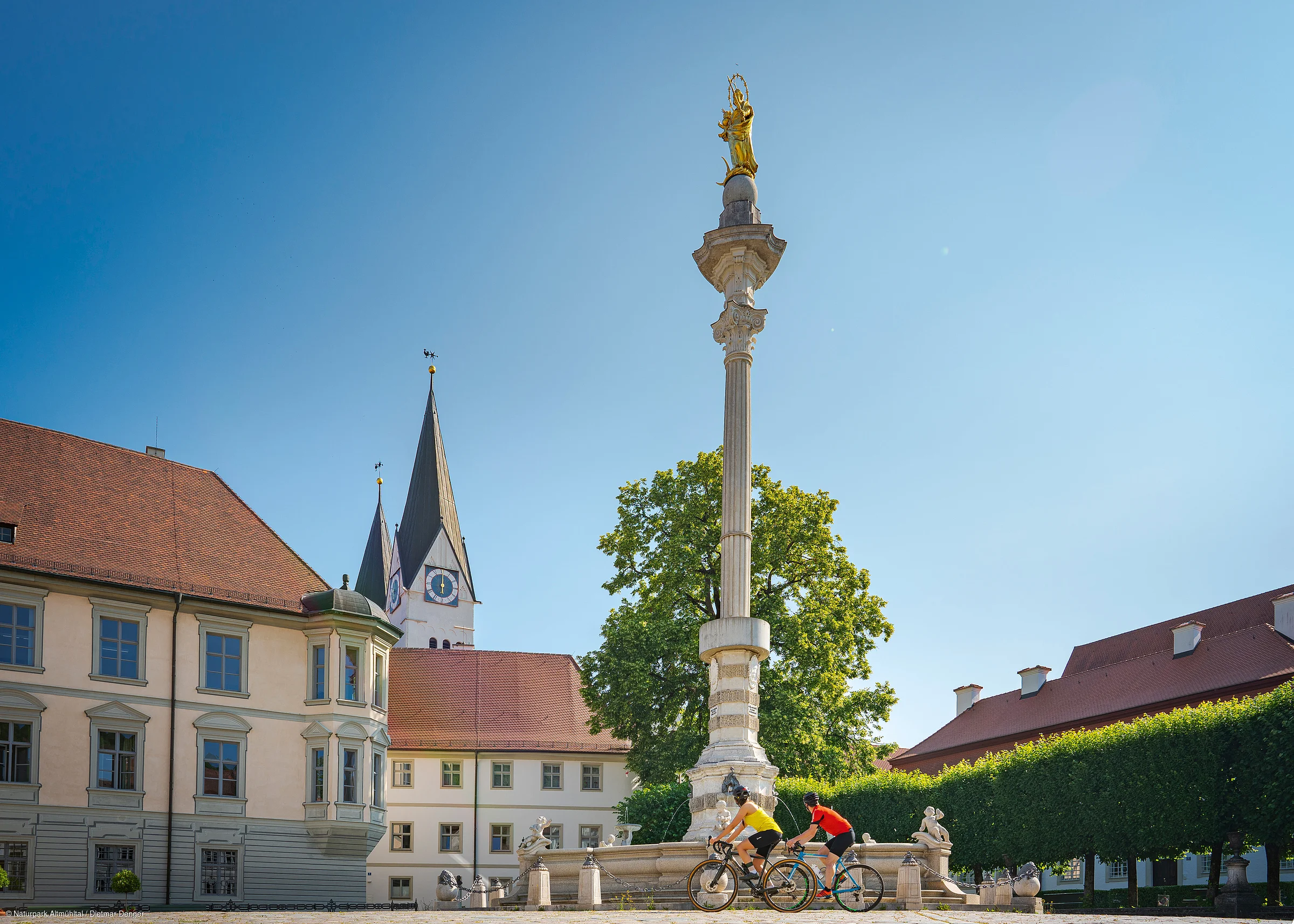 Zwei Radfahrer fahren an einer Mariensäule vorbei, im Hintergrund eine Kirche und Gebäude.