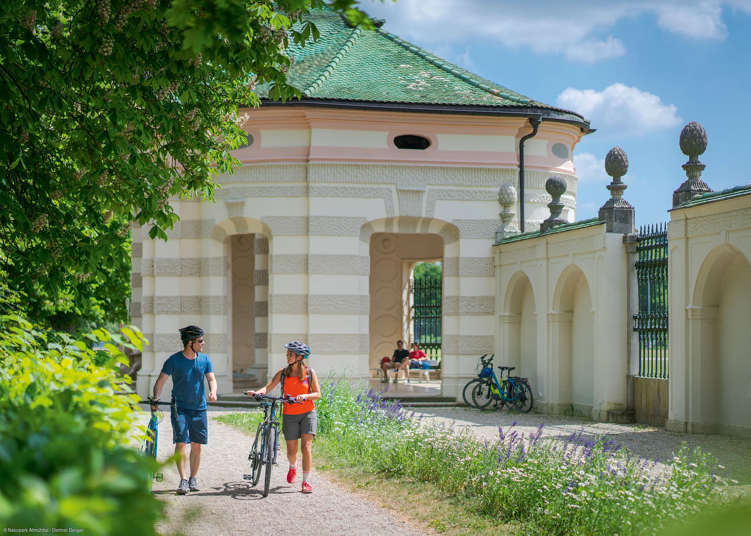 Zwei Personen mit Fahrrädern gehen auf einem Weg neben einem historischen Gebäude mit grüner Kuppel.
