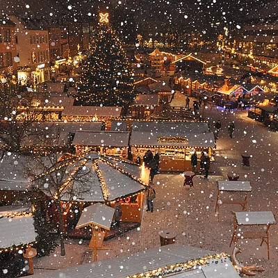 Verschneiter Weihnachtsmarkt mit beleuchteten Ständen und einem großen Weihnachtsbaum im Hintergrund.