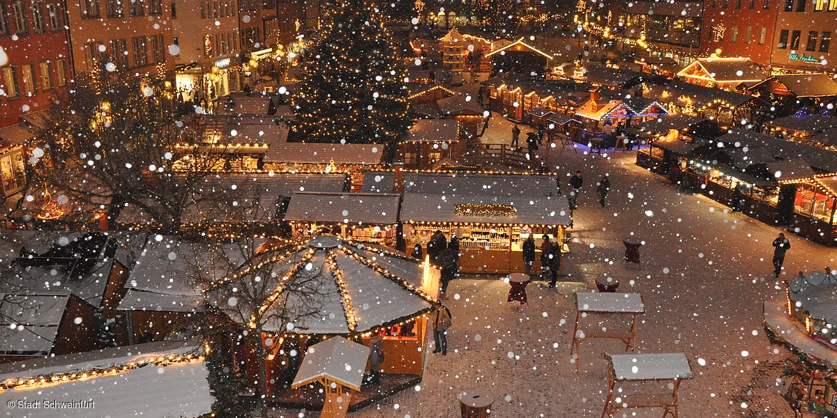 Verschneiter Weihnachtsmarkt mit beleuchteten Ständen und einem großen Weihnachtsbaum im Hintergrund.