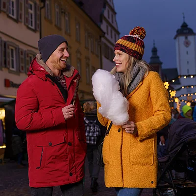 Zwei Personen in Winterkleidung essen Süßigkeiten auf einem festlich beleuchteten Weihnachtsmarkt.
