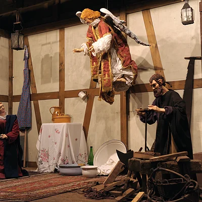 Krippenszene mit Figuren in einem Raum mit Holzwänden, ein schwebender Engel, ein Mann und eine Frau, Haushaltsgegenstände.