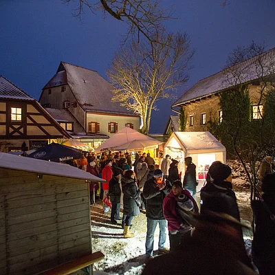 Menschen auf einem Weihnachtsmarkt mit Ständen und Fachwerkhäusern im Hintergrund bei Nacht.