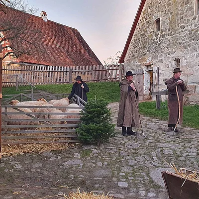 Drei Personen in historischen Kostümen mit Schafen auf einem gepflasterten Hof vor alten Gebäuden spielen Theater.