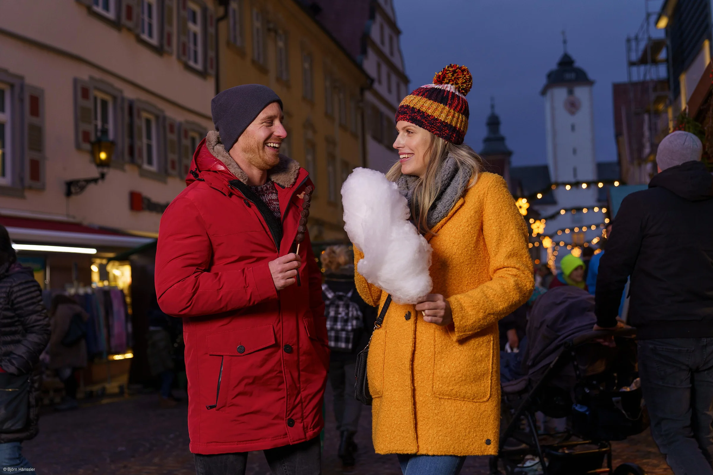 Zwei Personen in Winterkleidung essen Süßigkeiten auf einem festlich beleuchteten Weihnachtsmarkt.