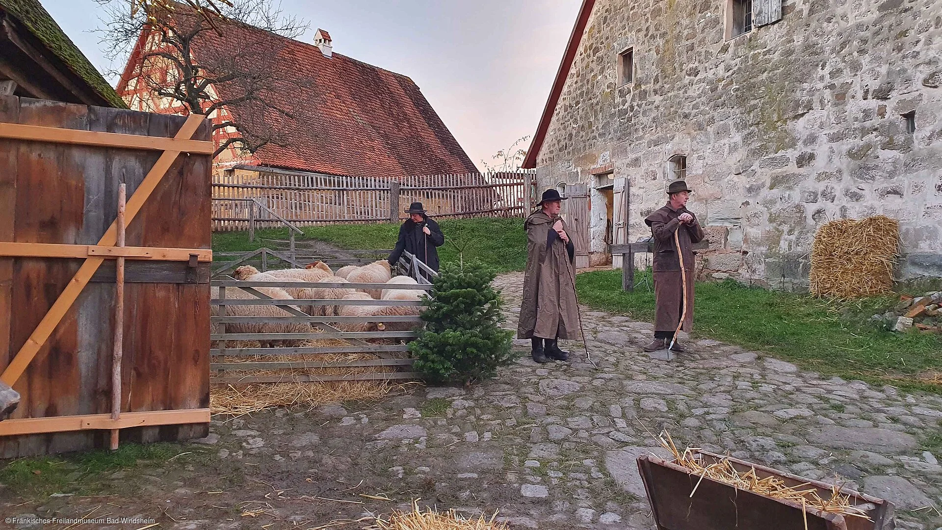 Drei Personen in historischen Kostümen mit Schafen auf einem gepflasterten Hof vor alten Gebäuden spielen Theater.