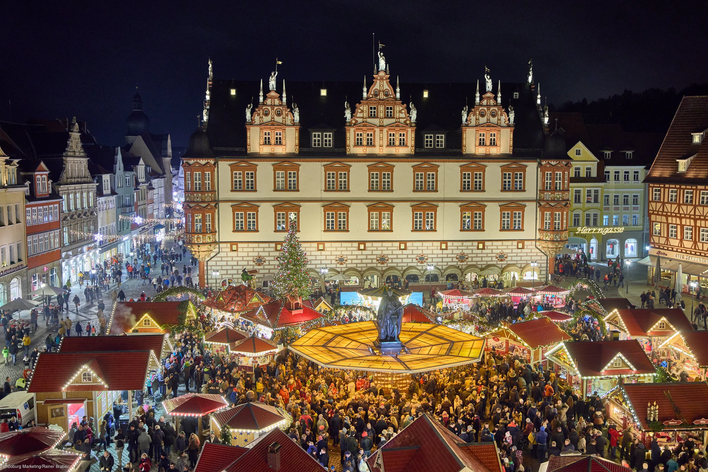 Weihnachtsmarkt mit beleuchteten Buden und einem großen Weihnachtsbaum vor einem historischen Gebäude bei Nacht.