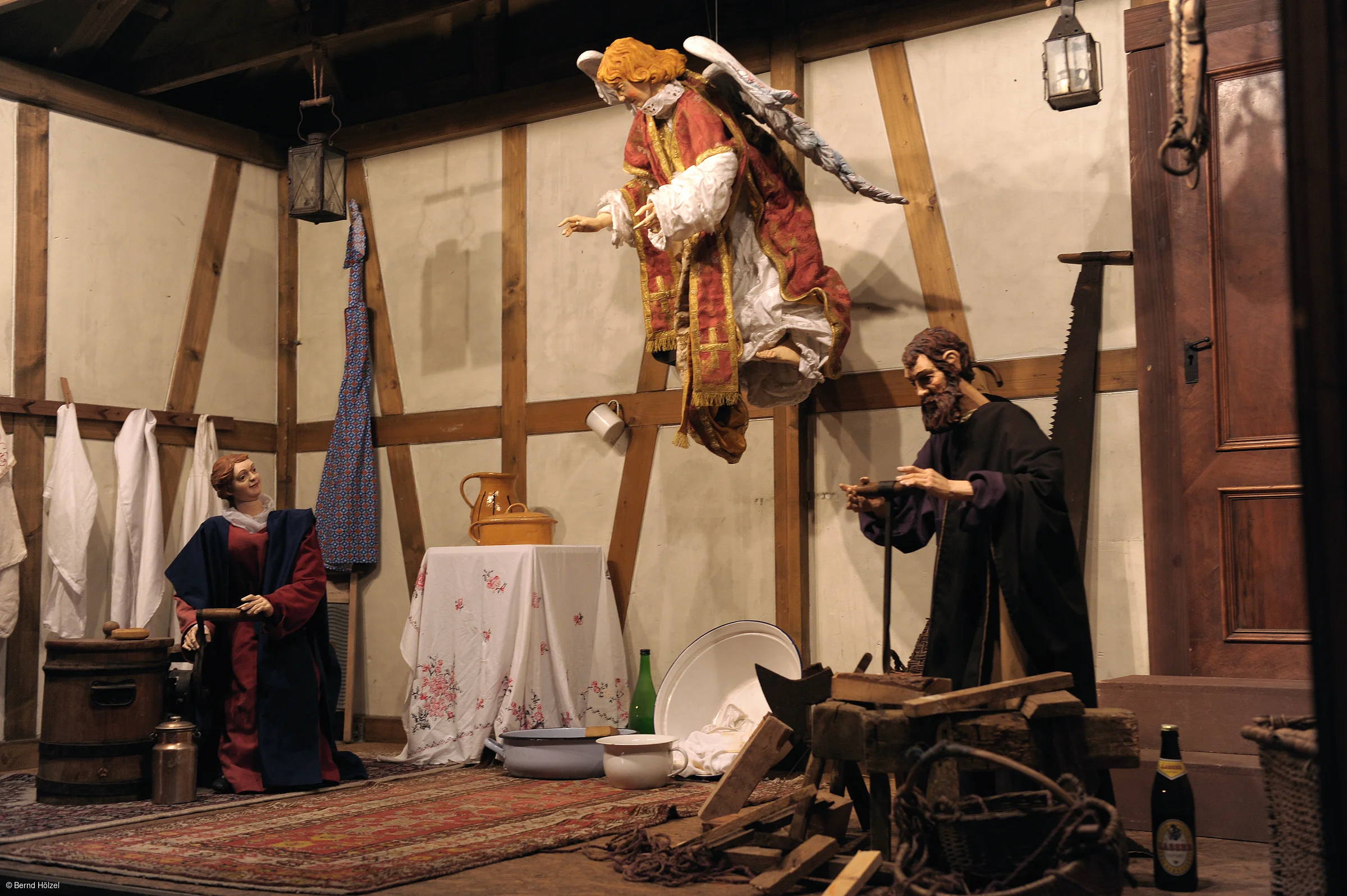 Krippenszene mit Figuren in einem Raum mit Holzwänden, ein schwebender Engel, ein Mann und eine Frau, Haushaltsgegenstände.