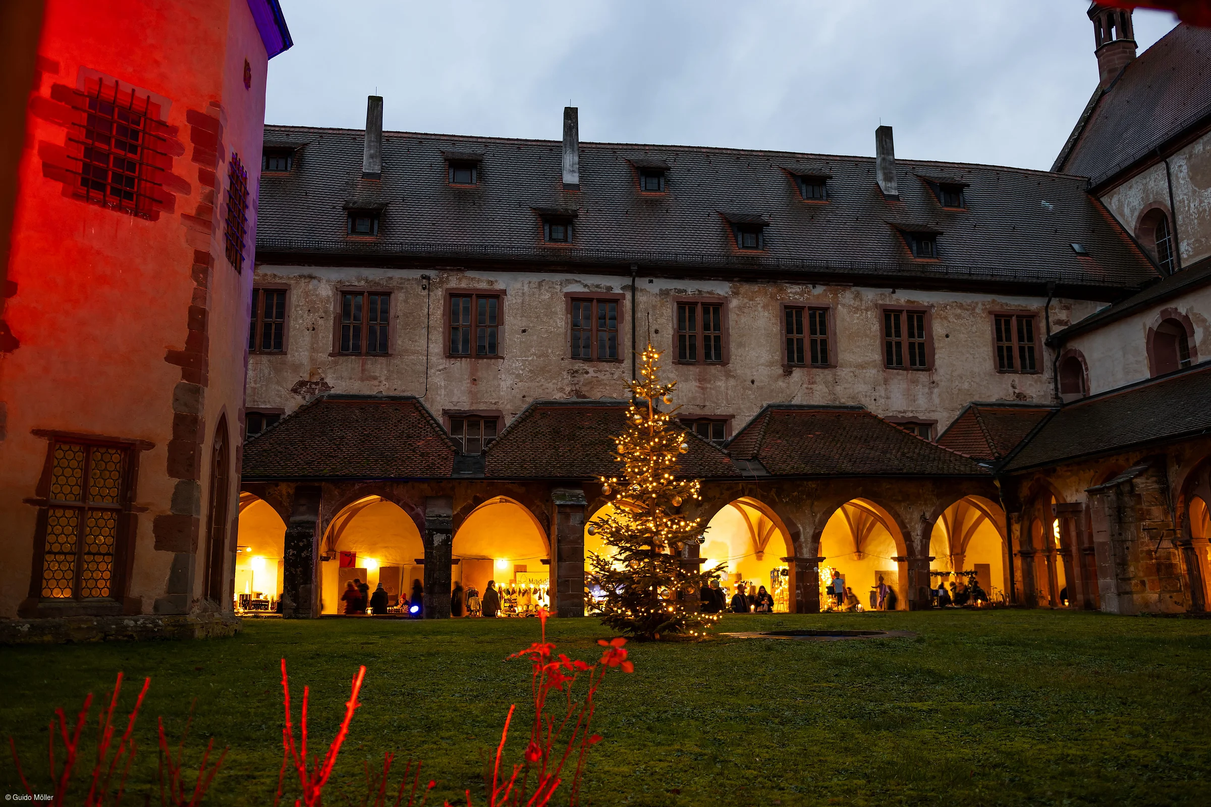 Historisches Klostergebäude mit beleuchtetem Weihnachtsbaum im Innenhof, umgeben von Arkaden und beleuchteten Fenstern.