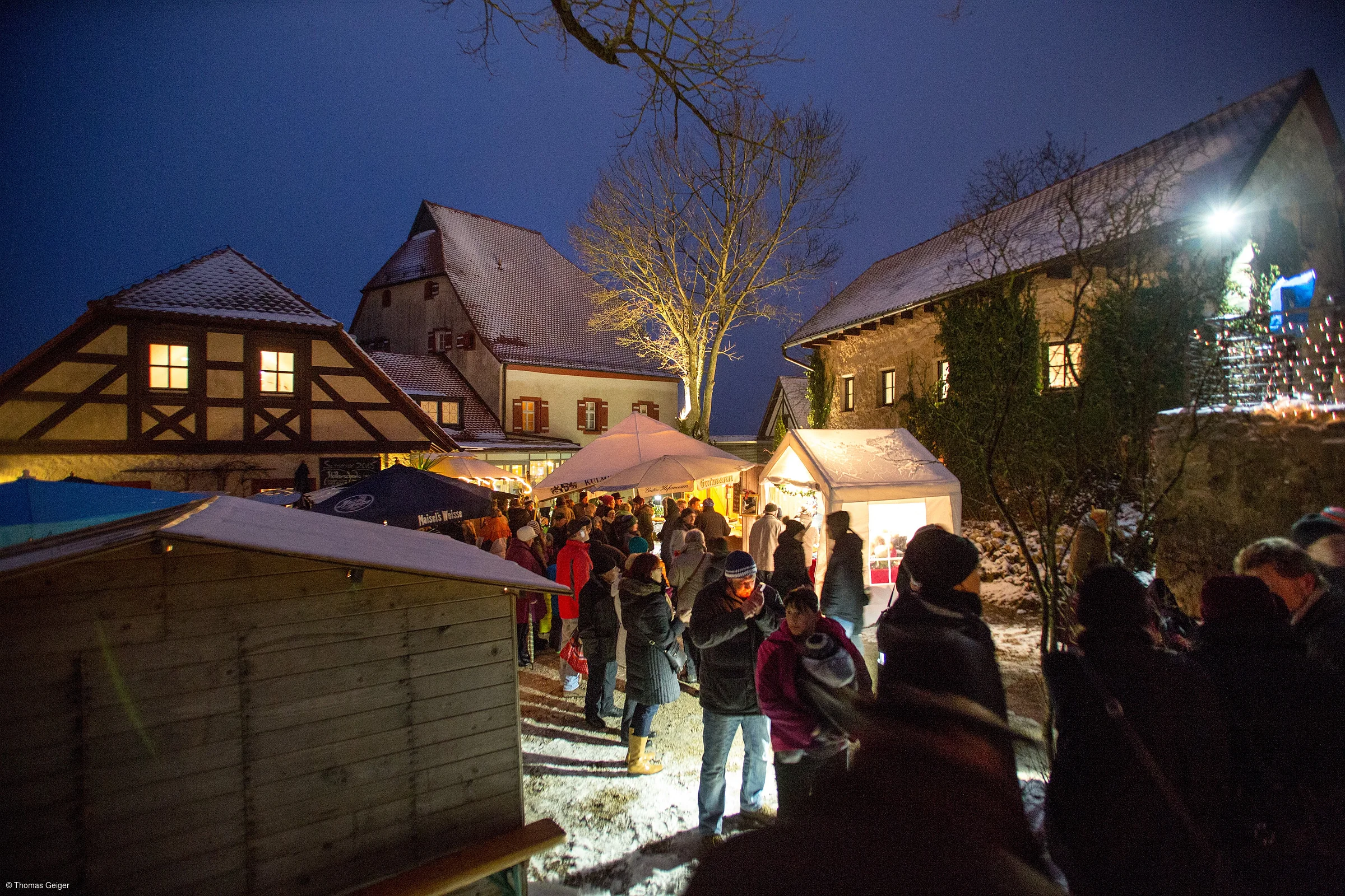 Menschen auf einem Weihnachtsmarkt mit Ständen und Fachwerkhäusern im Hintergrund bei Nacht.