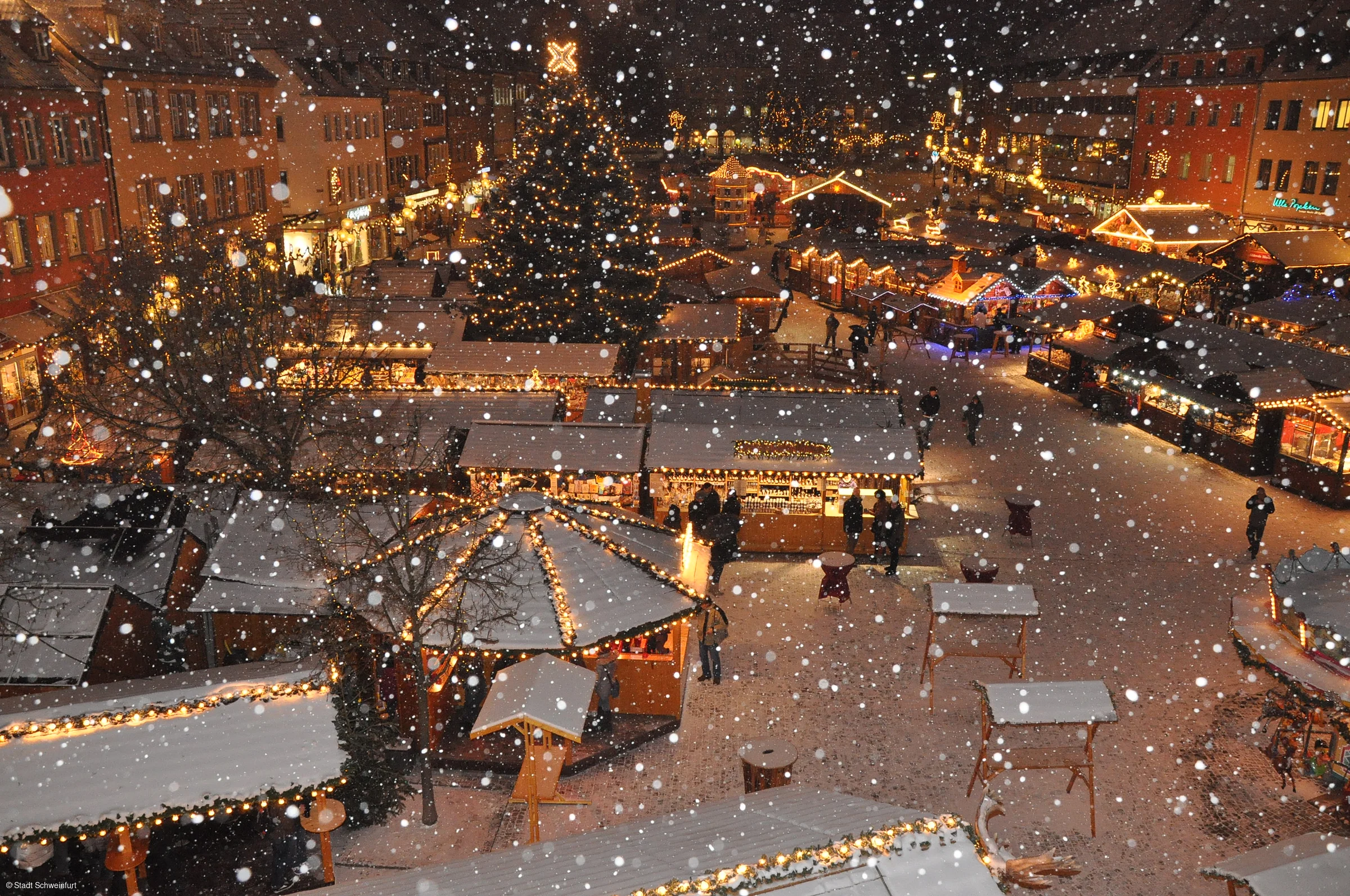 Verschneiter Weihnachtsmarkt mit beleuchteten Ständen und einem großen Weihnachtsbaum im Hintergrund.