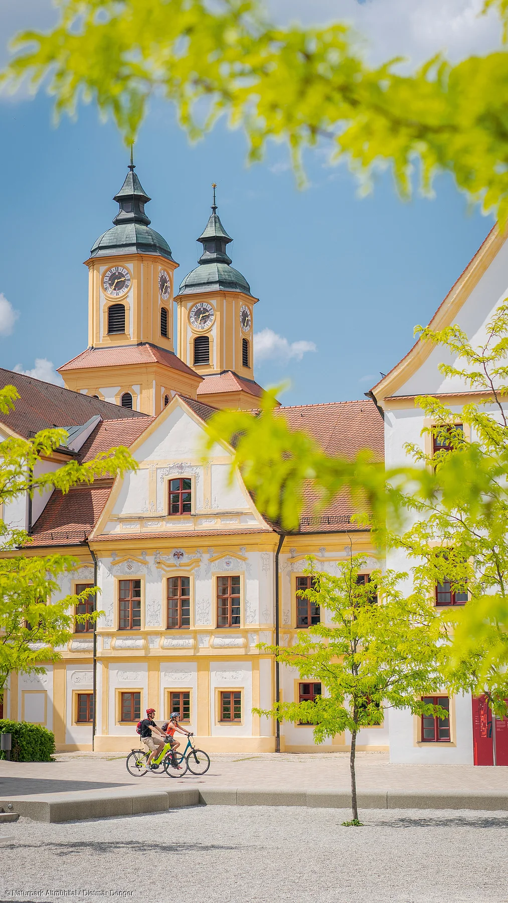 Zwei Personen fahren auf Fahrrädern vor einem historischen Gebäude mit zwei Kirchtürmen.