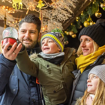 Familie auf einem Weihnachtsmarkt, hält eine Schneekugel. Hintergrund mit Lichtern und Dekorationen.