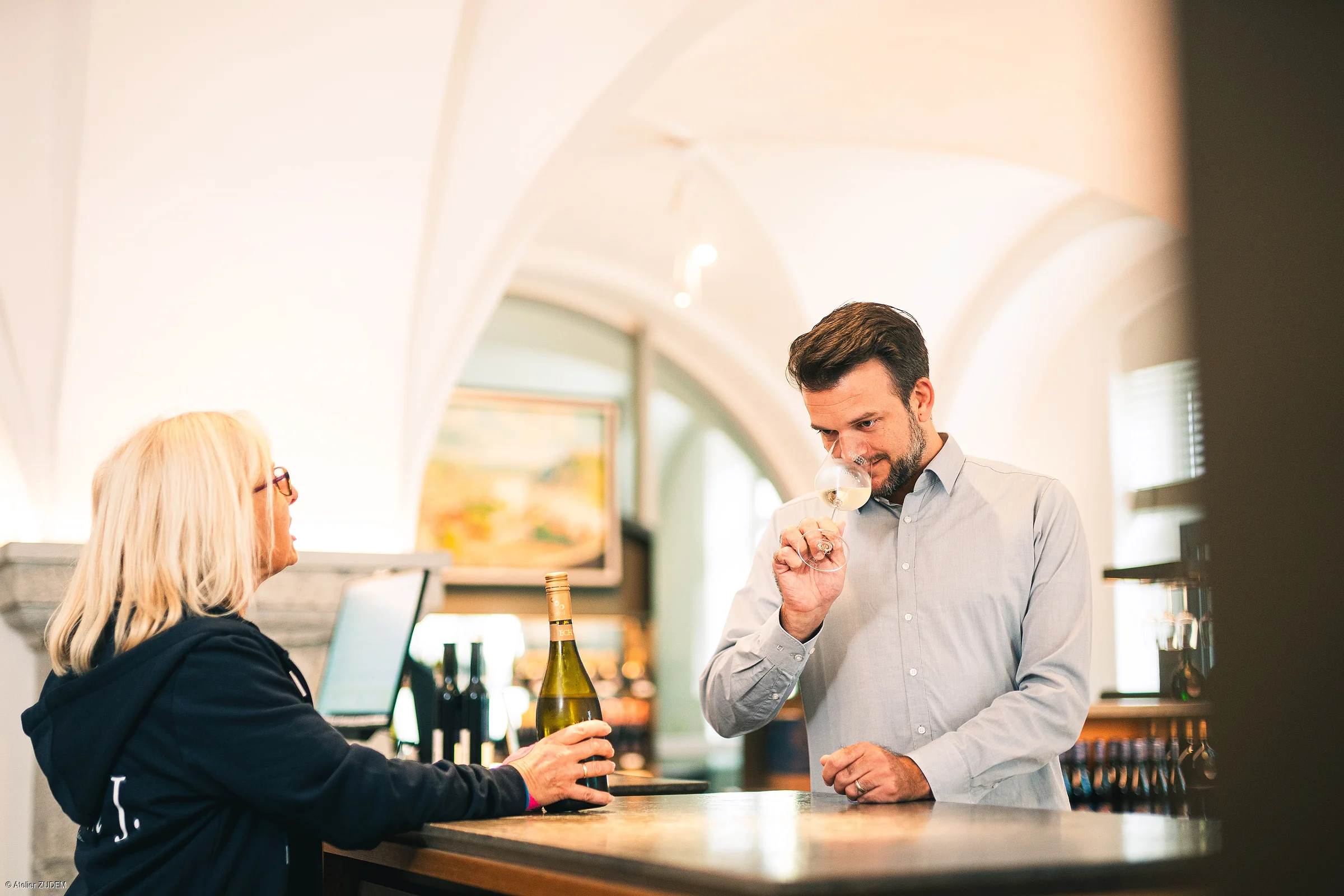 Mann riecht an Weinglas, Frau hält Weinflasche an Theke in hellem Raum mit gewölbter Decke
