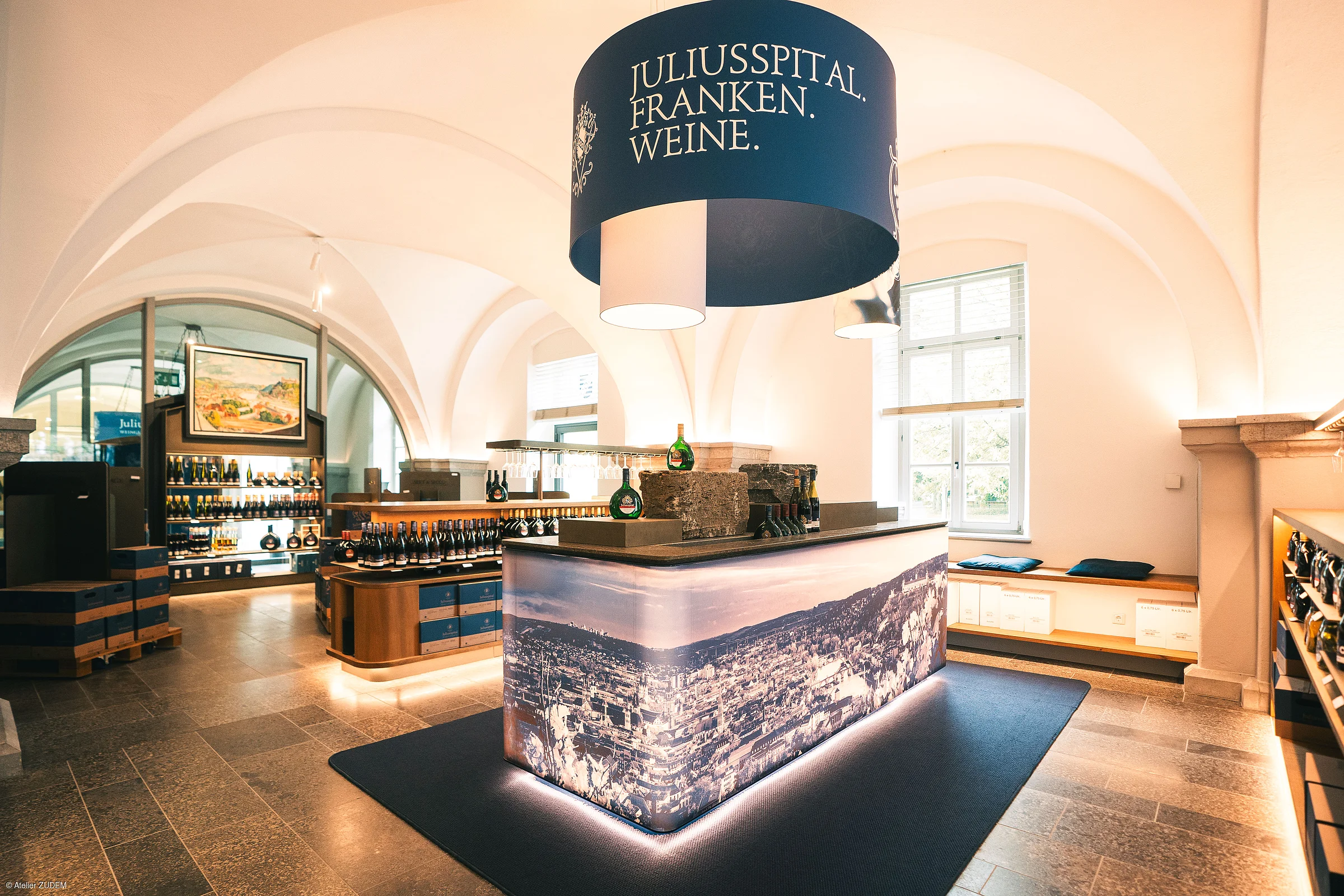 Innenraum eines Verkaufsraums mit Weinflaschen und einem beleuchteten Tresen unter einem runden Schild mit der Aufschrift „Juliusspital Franken Weine“