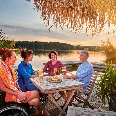 Vier Personen sitzen an einem Holztisch auf einem Steg am See bei Sonnenuntergang, eine Person im Rollstuhl.