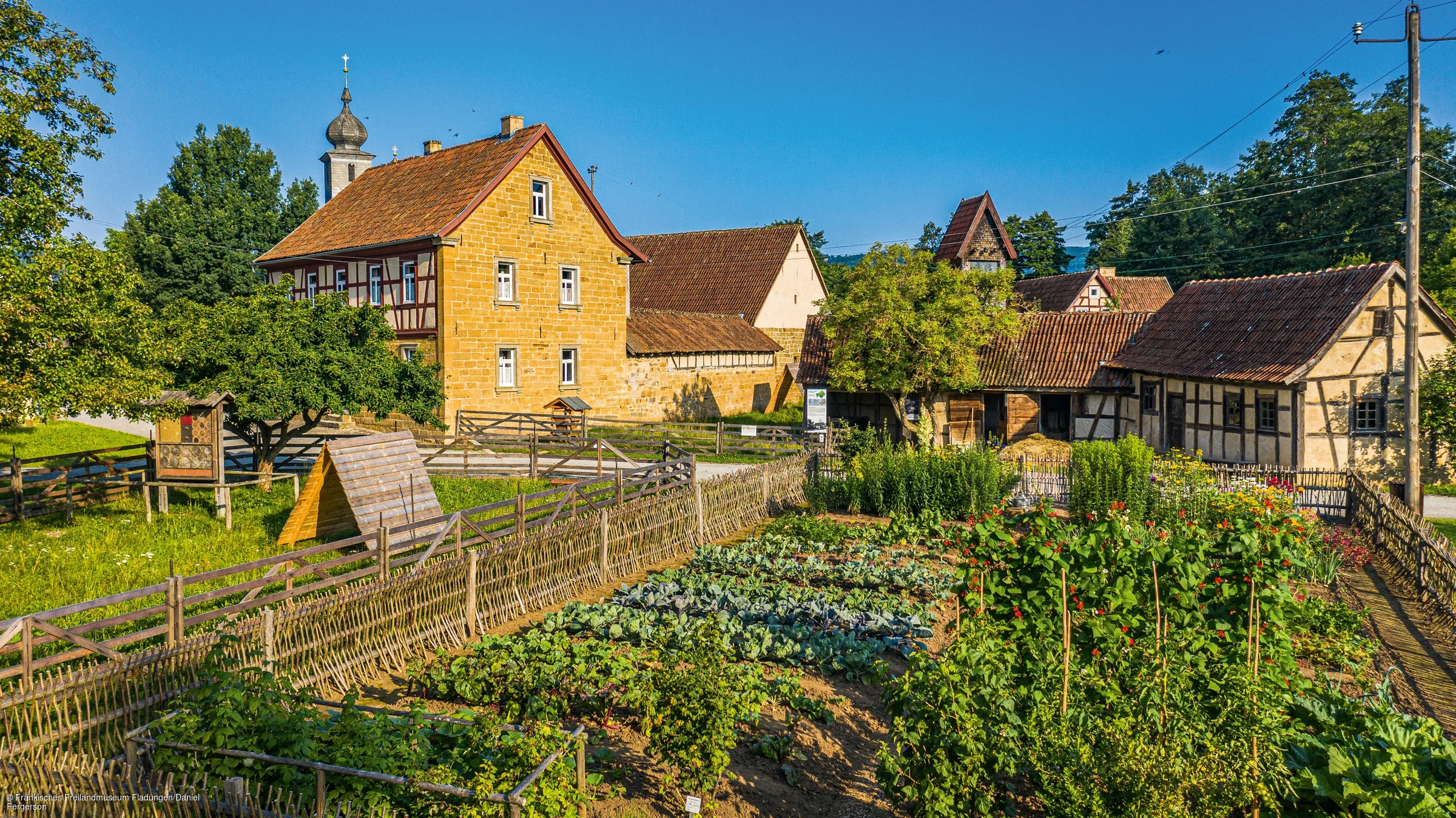 Bauernhof mit Gemüsegarten, Holzzäunen und Fachwerkhäusern unter blauem Himmel an einem sonnigen Tag.