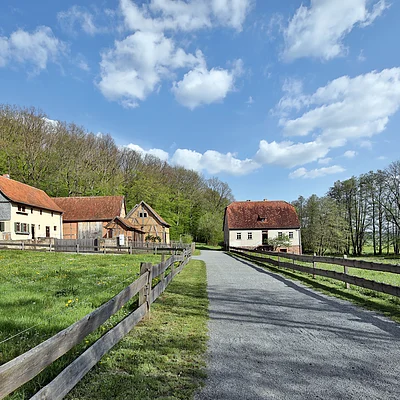Landstraße mit Holzzäunen führt zu mehreren Fachwerkhäusern vor bewaldetem Hügel unter blauem Himmel mit Wolken