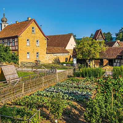 Bauernhof mit Gemüsegarten, Holzzäunen und Fachwerkhäusern unter blauem Himmel an einem sonnigen Tag.