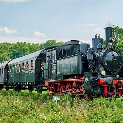 Dampflokomotive mit mehreren Waggons auf Gleisen in grüner Landschaft unter blauem Himmel