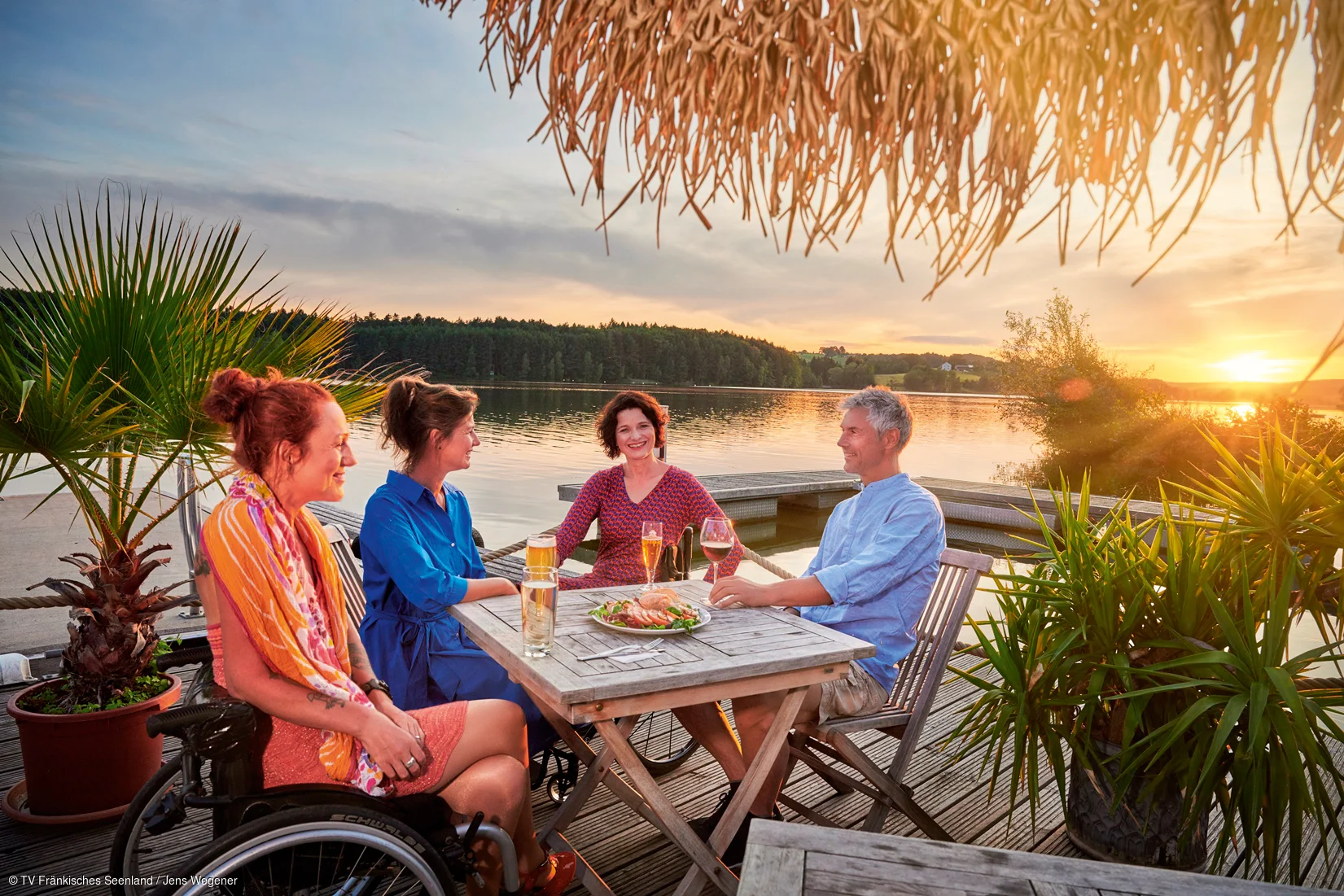 Vier Personen sitzen an einem Holztisch auf einem Steg am See bei Sonnenuntergang, eine Person im Rollstuhl.