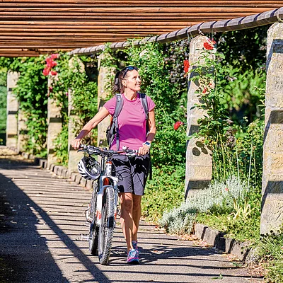 Frau in Sportkleidung schiebt Fahrrad unter Pergola mit Rosen und Säulen entlang eines Weges.