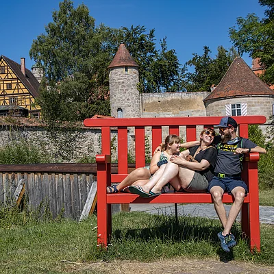 Drei Personen sitzen lachend auf einer übergroßen roten Bank vor einer historischen Stadtmauer mit Türmen.