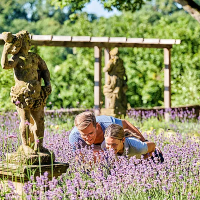 Zwei Personen riechen an lila Lavendelblüten in einem Garten mit Steinfiguren und Bäumen im Hintergrund.