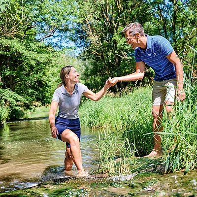 Frau steigt aus Bach mit Hilfe von Mann, umgeben von grünen Bäumen und Gras an sonnigem Tag.