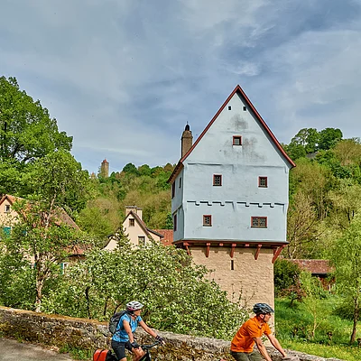 Zwei Radfahrer fahren auf einer Straße an einem historischen Haus mit Fachwerk und grüner Landschaft vorbei.