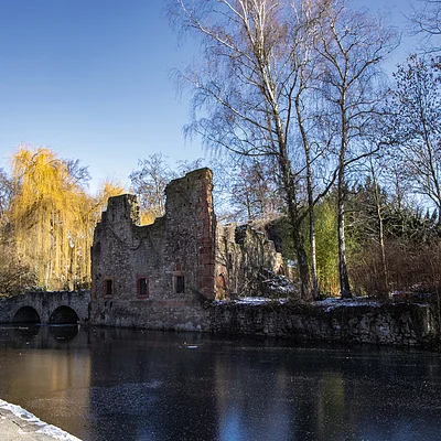 Ruine eines Steingebäudes an einem zugefrorenen Teich, umgeben von kahlen Bäumen und Winterlandschaft.