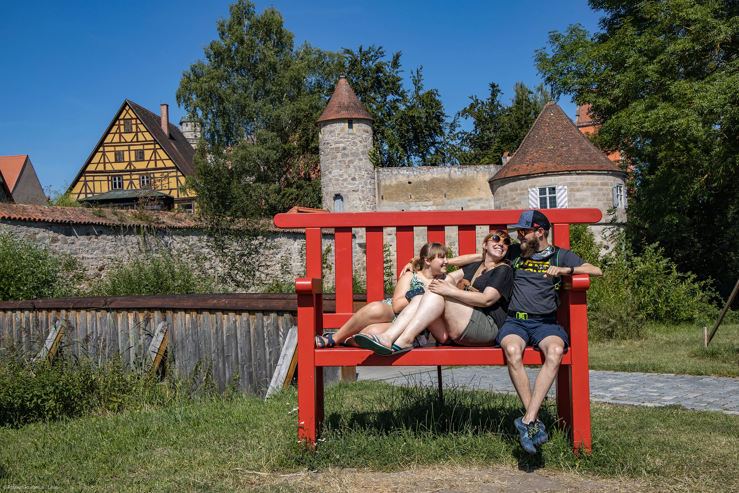 Drei Personen sitzen lachend auf einer übergroßen roten Bank vor einer historischen Stadtmauer mit Türmen.