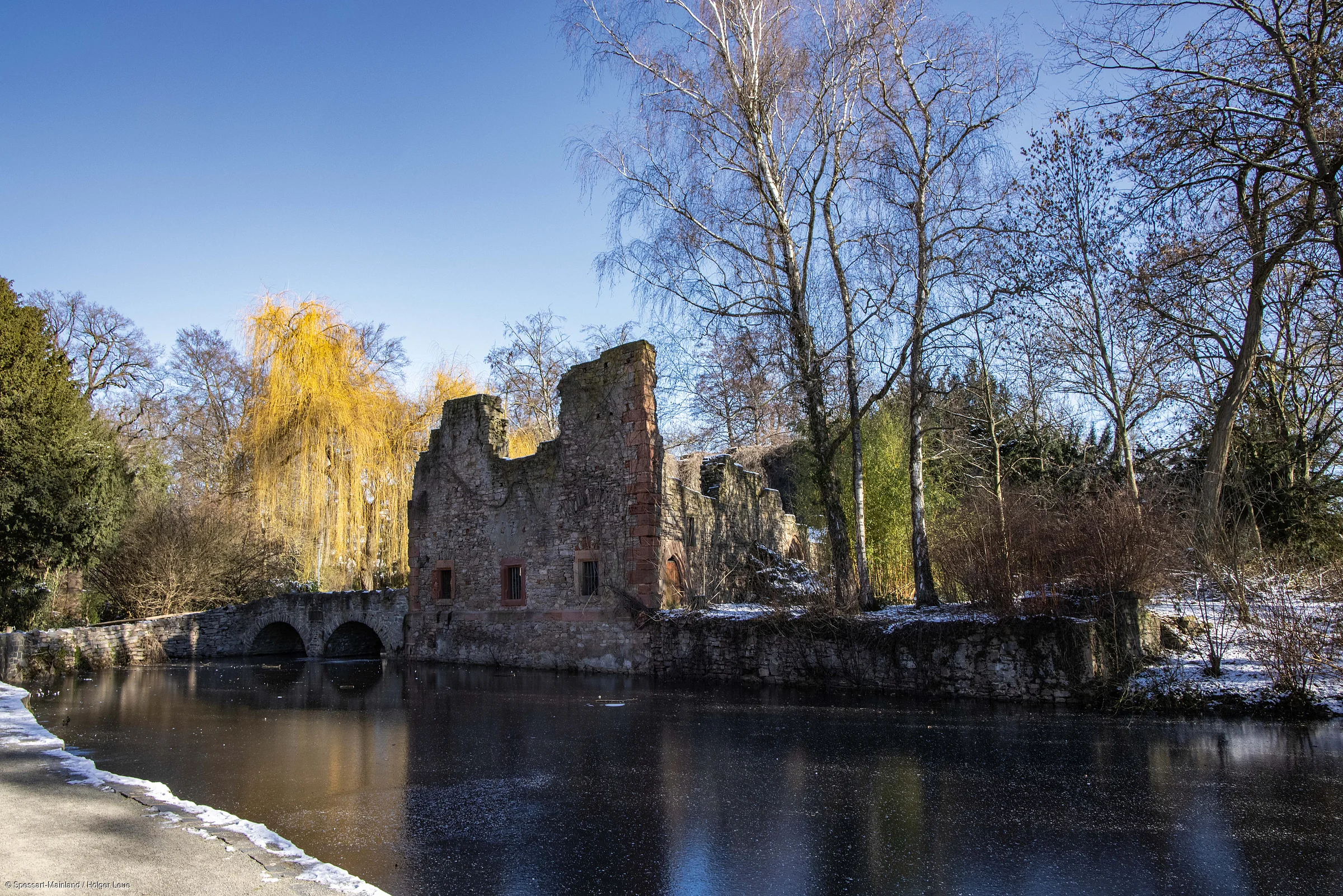 Ruine eines Steingebäudes an einem zugefrorenen Teich, umgeben von kahlen Bäumen und Winterlandschaft.