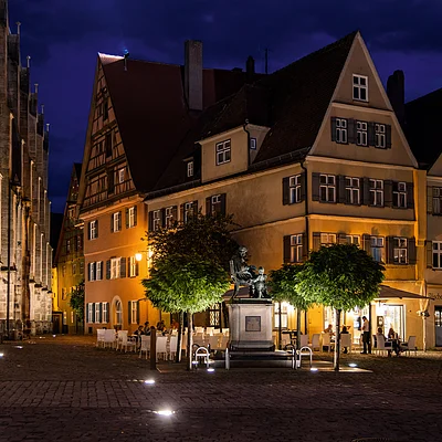 Nächtliche Altstadt mit beleuchtetem Fachwerkhaus, Bäumen und Statue auf gepflastertem Platz