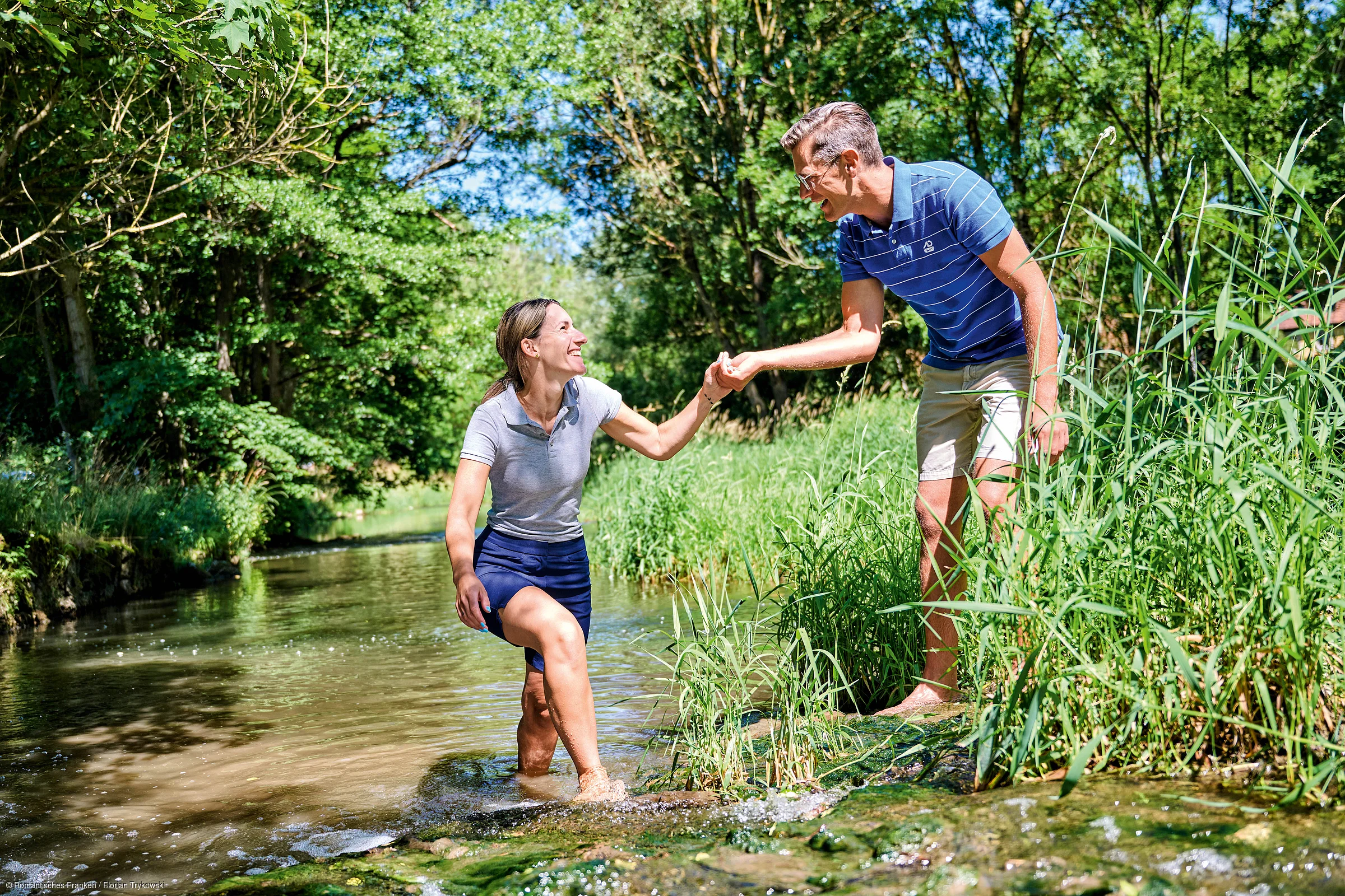Frau steigt aus Bach mit Hilfe von Mann, umgeben von grünen Bäumen und Gras an sonnigem Tag.