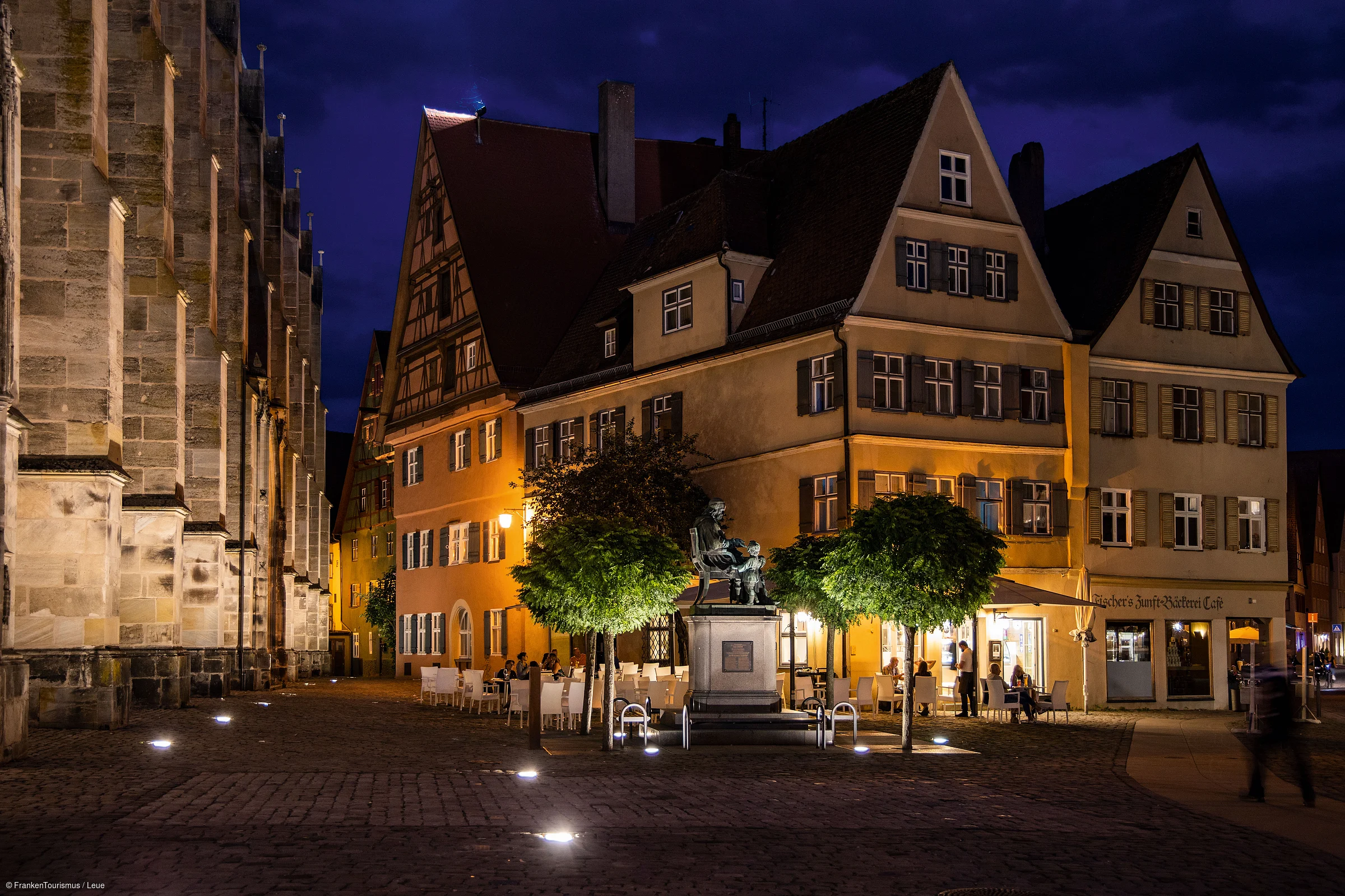 Nächtliche Altstadt mit beleuchtetem Fachwerkhaus, Bäumen und Statue auf gepflastertem Platz