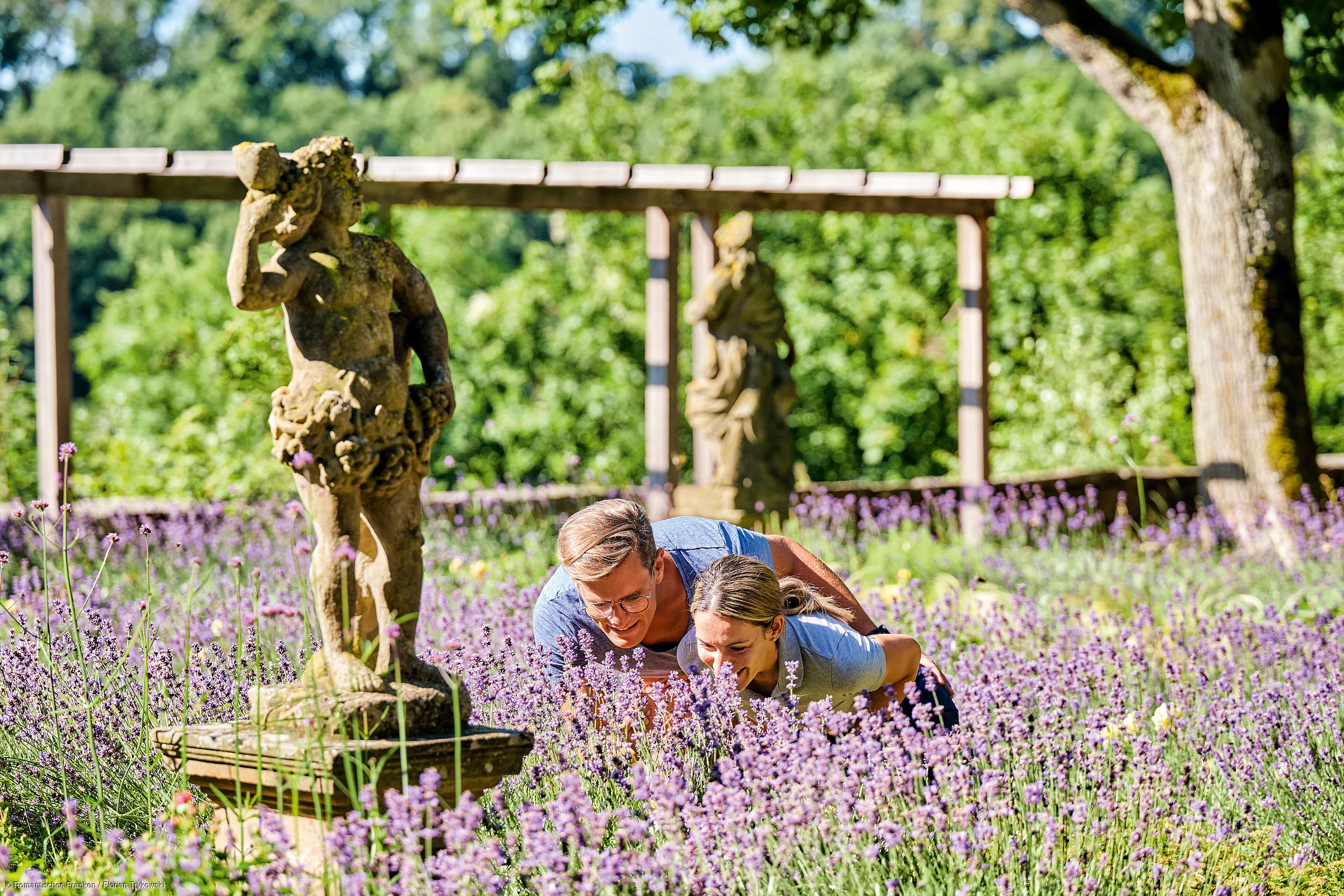 Zwei Personen riechen an lila Lavendelblüten in einem Garten mit Steinfiguren und Bäumen im Hintergrund.