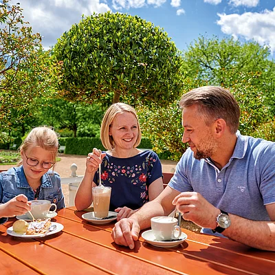 Familie mit Kind sitzt an Holztisch im Garten und trinkt Kaffee bei Sonnenschein