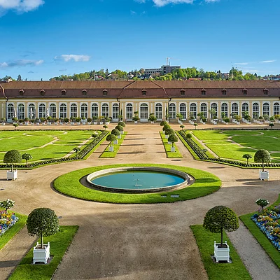 Barocker Garten mit symmetrischen Wegen, Blumenbeeten, Büschen und einem großen Gebäude im Hintergrund bei blauem Himmel.