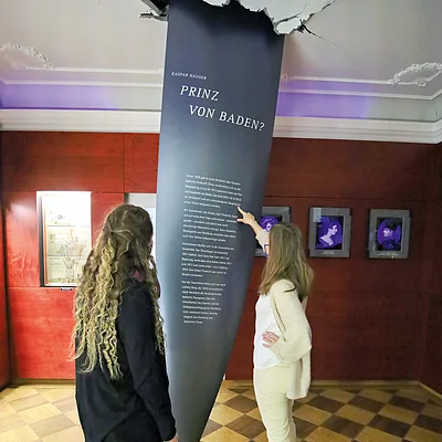 Zwei Frauen betrachten eine Museumsausstellung mit Texttafel, die aus der Decke zu brechen scheint.