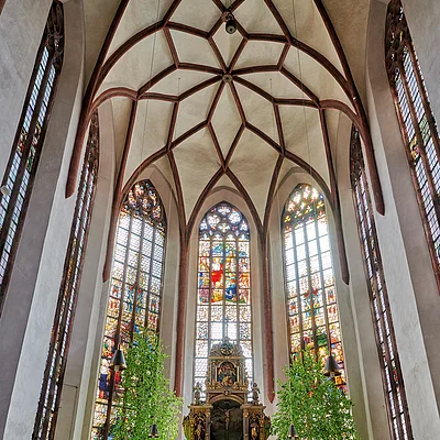 Innenraum einer Kirche mit hohen Buntglasfenstern, Holzbänken und einem Altar mit zwei grünen Bäumen.