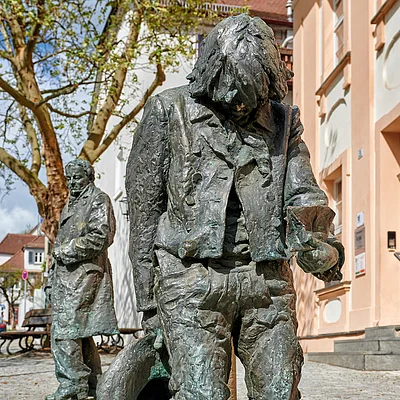 Bronzestatue eines Mannes mit Hut und Becher, im Hintergrund weitere Statue und Gebäude in einer Fußgängerzone.