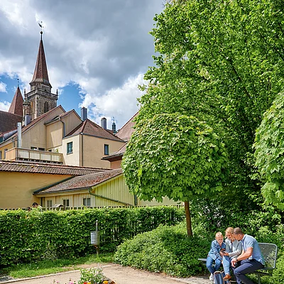 Familie mit Kind sitzt auf Parkbank im Garten, im Hintergrund Häuser und Kirchturm bei bewölktem Himmel.